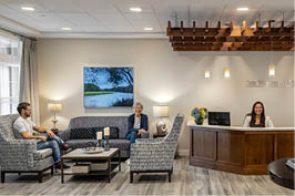 A woman is sitting at a desk in a lobby, with a man sitting on a couch nearby. There are two vases on the desk, and a potted plant is also present in the room. The setting appears to be a hotel lobby, with a TV mounted on the wall. AI generated content